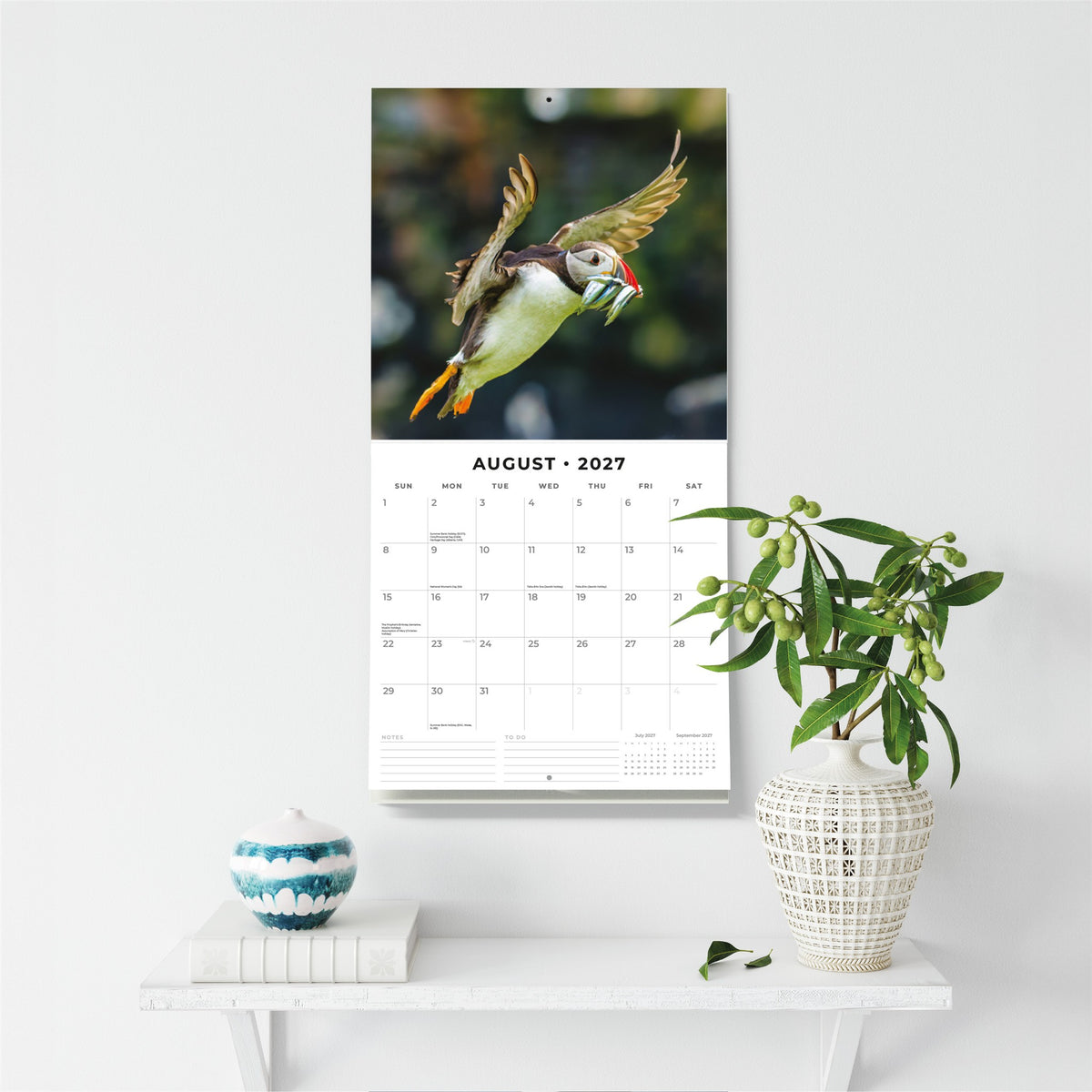 A wall-mounted calendar page featuring a photograph of a puffin carrying an fish, displayed above a monthly calendar grid for August 2027. The calendar hangs on a white wall above a small white shelf styled with a blue-and-white ceramic pot and a white vase with green leaves.